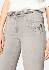 Vaqueros de mezclilla gris claro con adornos decorativos plateados y multicolores cerca de los bolsillos y un cinturón metálico a juego.