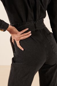 Pantalon en denim noir avec une taille haute, des poches latérales et une finition texturée. Une main repose sur la taille, mettant en valeur le design.