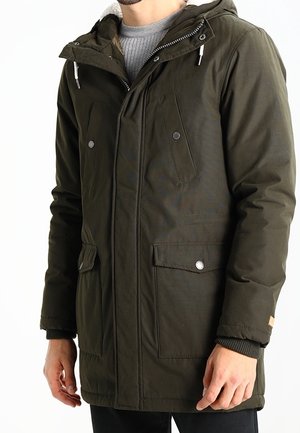 Uomo che indossa un parka con cappuccio verde oliva con tasche anteriori con bottoni e zip e polsini a coste sopra un maglione grigio.