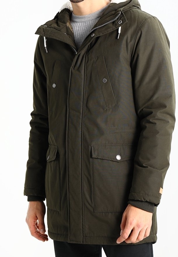 Homme portant une parka à capuche vert olive avec des poches avant à boutons et fermeture éclair et des poignets côtelés, par-dessus un pull gris.