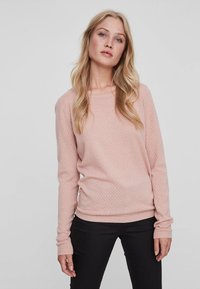 Lys rosa teksturert genser med lange ermer og rund hals, kombinert med svarte bukser. Materialet ser mykt og strikket ut.