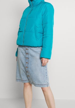 Teal puffer jacket zipped up, worn with knee-length light denim button-front skirt on a person standing against a plain wall.