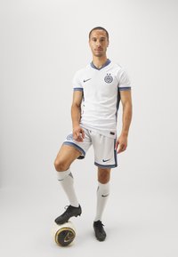 Witte voetbalkleding met blauwe accenten, voorzien van een v-hals, een logo op de borst en gestreepte mouwen. Model staat op een zwart-witte voetbal.