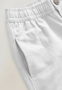 Pantalons en lin gris clair avec une taille élastique, présentant un détail de bouton et une texture subtile, montrés en gros plan.