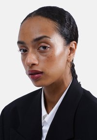 Junge Frau mit dunklem geflochtenem Haar, goldenen Creolen, Septumring, die einen schwarzen Blazer und ein weißes Hemd trägt und selbstbewusst in die Kamera schaut.