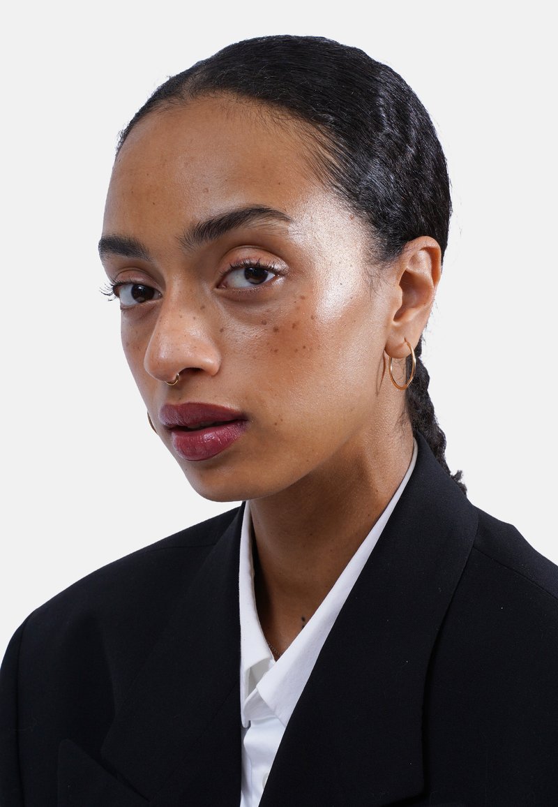 Junge Frau mit dunklem geflochtenem Haar, goldenen Creolen, Septumring, die einen schwarzen Blazer und ein weißes Hemd trägt und selbstbewusst in die Kamera schaut.