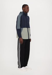 Vindjakke i marineblå, grå og lysegrønn farge, med hette og elastiske ermer, kombinert med svarte joggebukser med hvite sidestriper.