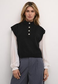 Black knit sleeveless top featuring a high neck and five silver buttons. Worn over a sheer white long-sleeve blouse with gathered cuffs.