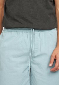 Lichtblauwe shorts van textiel met een elastische tailleband en trekkoord, gecombineerd met een donkergrijs shirt. Handen rusten op de shorts.