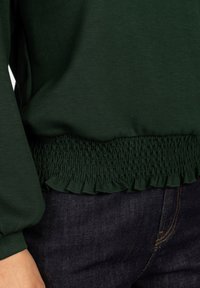 Groene sweatshirt met een geschulpte ruffle onderrand. Gemaakt van zachte stof, met lange mouwen en een relaxte pasvorm, gecombineerd met donkere jeans.