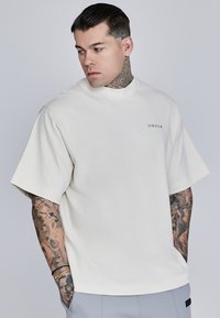 Valge puuvillane T-särk kõrge kaelusega ja lühikeste varrukatega, millel on rinnal väike must logo ja lõdvelt fit.