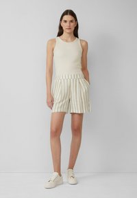 Beige Tank-Top und gestreifte Leinen-Shorts in Creme und Hellbraun. Die Shorts haben Taschen und einen umgeschlagenen Bund. Weiße Sneakers runden das Outfit ab.