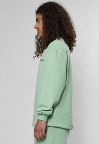Felicissimo verde chiaro in morbido tessuto, con maniche lunghe, vestibilità rilassata e polsini a coste. Design semplice con branding minimale.