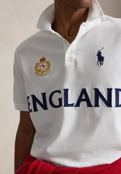 Άτομο που φοράει λευκό πόλο με το κείμενο «ENGLAND», ένα κεντημένο κόκκινο και χρυσό έμβλημα και ένα λογότυπο παίκτη πόλο σε σκούρο μπλε στο στήθος.