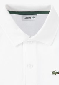 Λευκό πόλο μπλουζάκι από υφασμάτινη ύφανση, με πράσινη κολλάρο λεπτομέρεια και μικρό κεντημένο λογότυπο πράσινου κροκόδειλου στο στήθος.