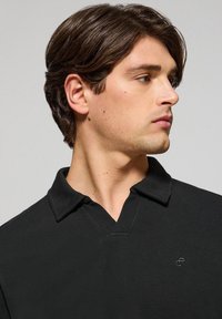 Polo negro con tejido texturizado, con escote en V y mangas cortas. Pequeño logo bordado en el lado izquierdo del pecho.