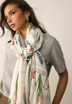 Junge Frau mit langen dunklen Haaren, die goldene Creolen trägt, ein graues Oberteil und einen großen weißen Schal mit bunten Blumenmustern um den Hals gebunden.