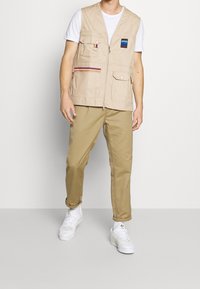 Beige ärmlös väst med flera fickor, dragkedja framtill, broderad logotyp och dekorativ rand, kombinerad med khakifärgade byxor och vita sneakers.
