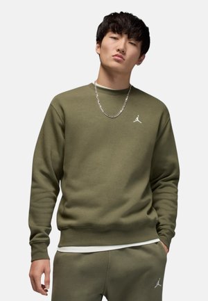 Giovane uomo con felpa Jordan verde oliva e pantaloni della tuta abbinati, in piedi con una mano in tasca contro uno sfondo semplice.