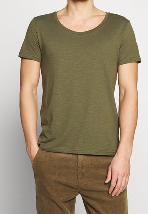 Personne portant un t-shirt vert olive à manches courtes et un pantalon en velours côtelé marron, une main dans la poche, sur un fond uni.