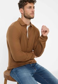 Bruine langemouwen polo met een button-down kraag, subtiele textuur en voor sluiting met knopen. Draag het met blauwe denim jeans.