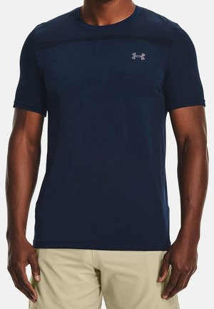 Mann trägt ein eng anliegendes marineblaues Under Armour Kurzarmshirt und beige Shorts, steht vor weißem Hintergrund.