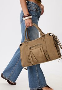 Borsa in suede beige con doppi manici, tasca frontale con zip e dettaglio in frange. Abbinata a jeans in denim blu a palazzo.