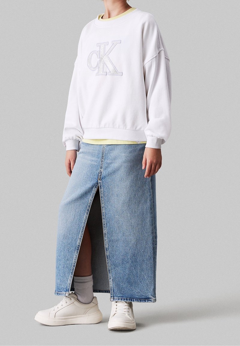 Personne portant un sweat-shirt blanc Calvin Klein, une jupe en denim bleu clair avec une fente à l'avant, des chaussettes blanches et des baskets blanches, debout devant un fond gris.