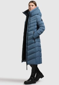 Garšs zils pūkains mētelis ar augstu apkakli, vertikālu quilted dizainu, ar rāvējslēdzēju un sānu kabatām. Nēsāts ar melniem, elegantiem apaviem.