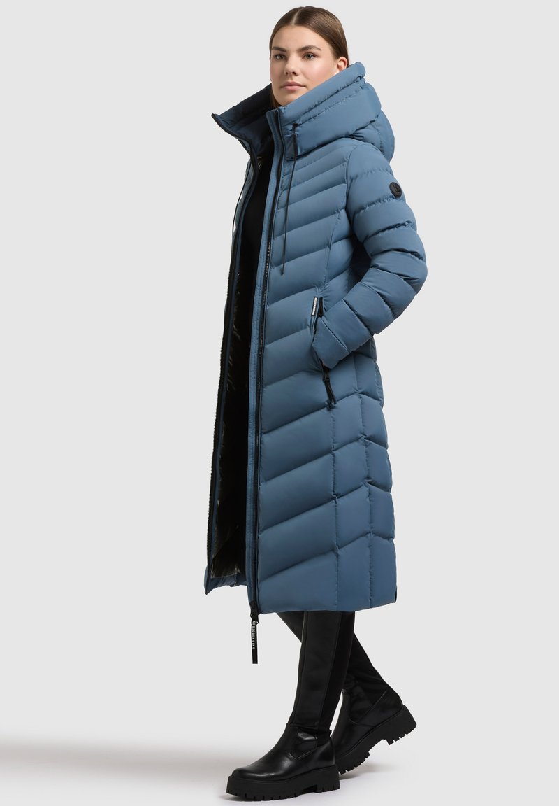 Garšs zils pūkains mētelis ar augstu apkakli, vertikālu quilted dizainu, ar rāvējslēdzēju un sānu kabatām. Nēsāts ar melniem, elegantiem apaviem.