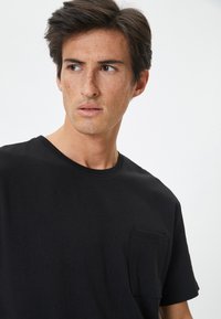 Camiseta de algodón negra con cuello redondo, mangas cortas y un bolsillo en el pecho. La tela parece suave y tiene un corte relajado.