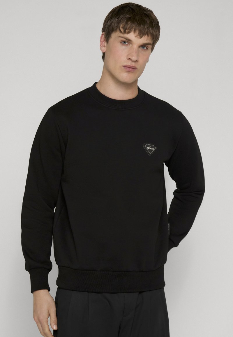 Jeune homme aux cheveux courts portant un pull noir à manches longues avec un petit emblème en forme de losange sur la poitrine.