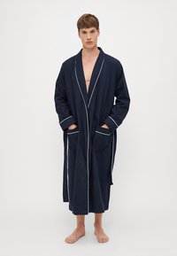 Marineblå badekåpe laget av myk stoff, med sjal-krage, hvite kanter, lange ermer og to frontlommer. Gulvlengde design.