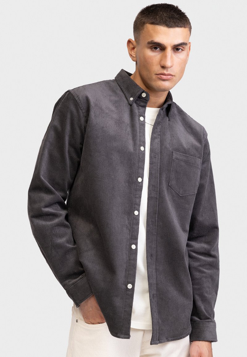 Graues Cordhemd mit Button-Down-Kragen, Brusttasche, strukturiertem Muster und weißen Knöpfen, getragen über einem weißen T-Shirt.