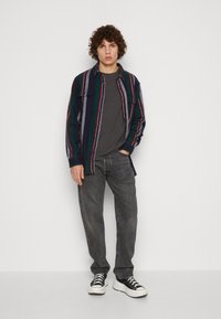 Camisa de franela en color marino con rayas verdes y burdeos, diseño de botones, dos bolsillos frontales, superpuesta sobre una camiseta gris, combinada con jeans grises.