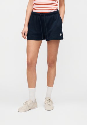 Person iført marineblå snor-shorts, hvite sokker, beige joggesko og en rødt- og hvitstripet topp, står foran en hvit bakgrunn.