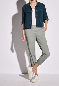 Chemise en daim bleu marine avec fermeture à boutons, deux poches poitrine ; pantalon tapered vert clair ; baskets blanches à lacets avec semelles jaunes.