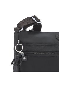 Schwarze Nylon-Tasche mit einem dunkelbraunen Stoffgurt, silberner Hardware, einer vorderen Reißverschlusstasche und einem angehängten schwarzen Gummianhänger.