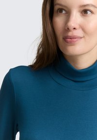 Frau mit heller Haut und braunen Haaren, die einen türkisfarbenen Rollkragenpullover trägt, leicht nach rechts blickend und mit einem neutralen Gesichtsausdruck.