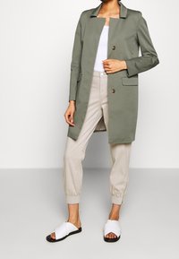 Olivově zelený dlouhý kabát s výstřihem, dvěma knoflíky a bočními kapsami, kombinovaný s béžovými kalhotami a bílými klouzavými sandály.