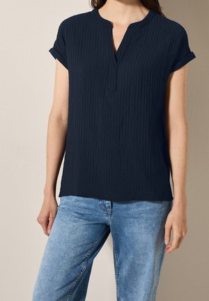 Granatowa bluzka z teksturowanej tkaniny, z kołnierzykiem w kształcie litery V i krótkimi, podwiniętymi rękawami, w połączeniu z jasnoniebieskimi dżinsami.
