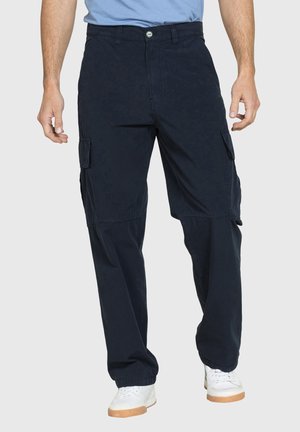 Uomo che indossa pantaloni cargo blu navy con tasche laterali e sneakers bianche, in piedi contro uno sfondo neutro.