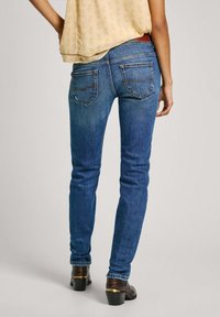 Modré džíny skinny s klasickými kapsami a středním pasem, doplněné hnědými botami na podpatku a žlutým vzorovaným topem.