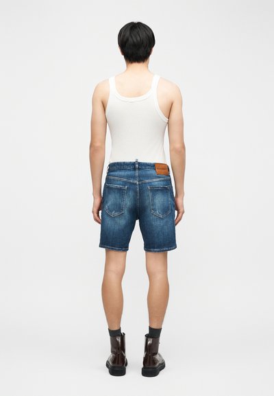 Homme de dos portant un débardeur blanc, un short en jean bleu, des chaussettes foncées et des bottines marron, debout sur un fond clair uni.