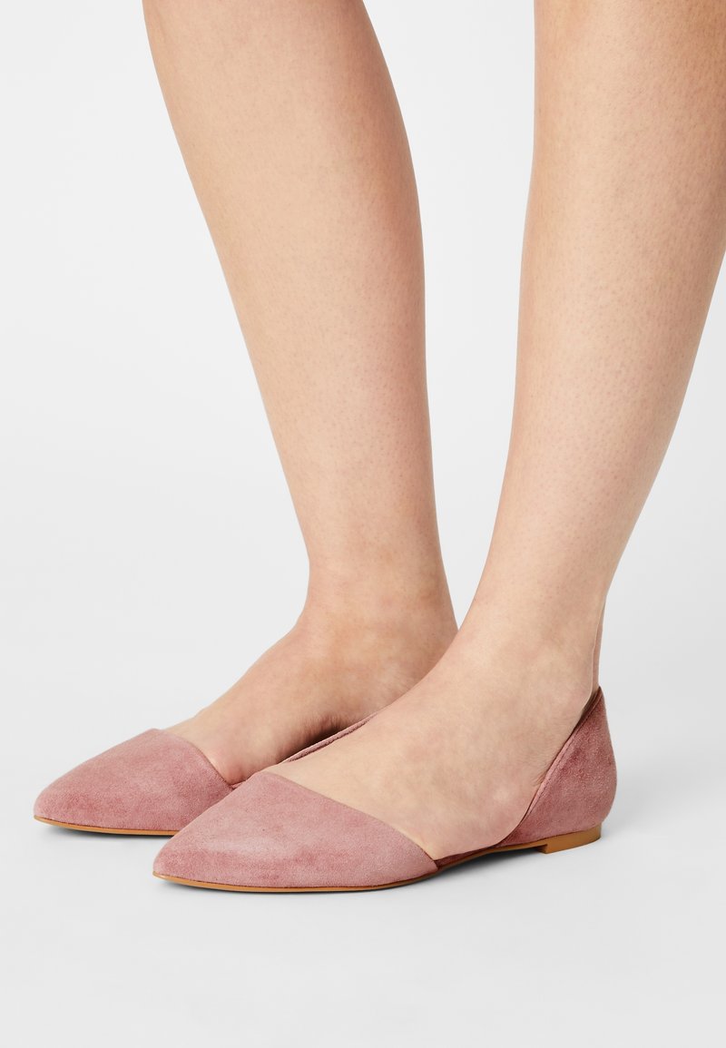 Jambes portant des chaussures plates en daim rose poudré à bout pointu avec de petits talons sur un fond clair uni.