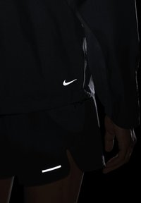 Musta urheilu takki ja shortsit, joissa on valkoiset Nike-logot ja heijastavat yksityiskohdat. Tekstuuroitu kangas kevyellä muotoilulla, slim fit.
