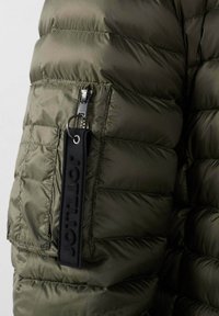 Giacca trapuntata verde oliva con tasca con zip e un portachiavi nero con il marchio attaccato al tirante della zip.