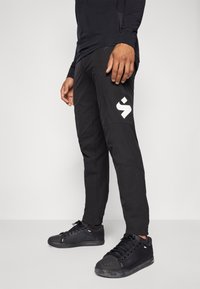 Juodos tapered kelnės, pagamintos iš tamprios medžiagos, su dideliu baltu logotipu ant šlaunies, derinamos su juodomis sportbačiais.