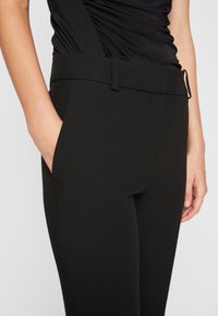 Pantalons noirs ajustés avec une texture lisse, poches latérales et une taille plate. Présente un détail de boucle discret sur le bord de la taille.