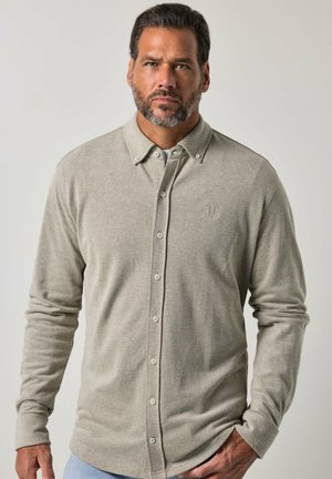 Mand med kort mørkt hår og skæg iført en lysebeige skjorte med knapper og lange ærmer mod en ensfarvet lys baggrund.